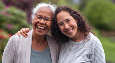 A older woman embraces her daughter as they happily walk through a park and enjoy their time together. The daughter is smiling at the camera.